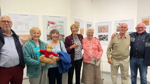 BBC Former internees smile at the camera as they stand in front of some of the displays at the exhibition