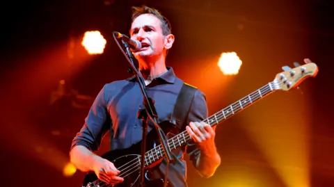 Getty Images Jim Glennie playing a black bass guitar on stage, while singing into a microphone.