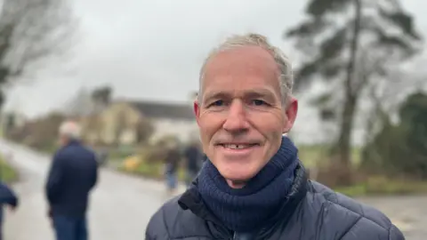 Joao Santos/LDRS A head and shoulders image of Darren Lewitt. He is wearing a navy blue jacket and a navy blue scarf. He is smiling and looking directly into the camera. The background is blurred but there appears to be a road, some trees and other people.