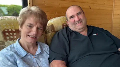 Marshall, a big man in a dark polo shirt, sits beside a woman in a pin-stripped shirt on a sofa. Behind them is a wood panelled interior wall. 