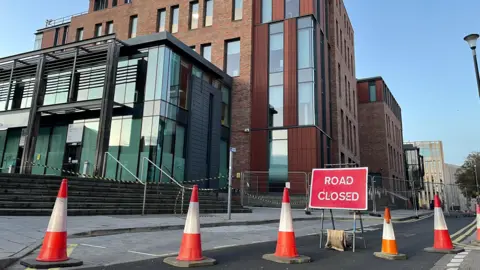 Traffic cones, temporary metal fences, black-and-yellow tape and a red 'Road Closed' sign outside Durham Passport Office.