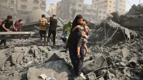 Woman holding a baby in the middle of ruins following an attack, with others comforting each other or looking through the rubble