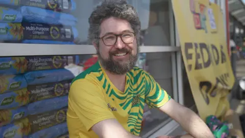 Hannah Karpel/ BBC A man wears a yellow top and sits in front of a supermarket smiling into the camera. He wears glasses and has grey hair and a grey beard.
