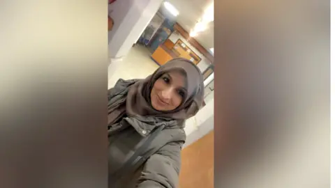 Shabnam Sabir - a young woman in a hijab taking a selfie from a jaunty angle. She is wearing a beige winter coat and smiling into the camera.