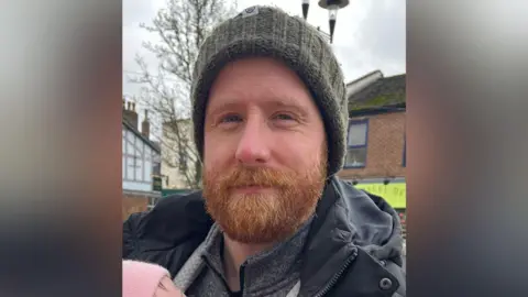Philip Gilligan as a ginger beard and is wearing a grey woollen hat a navy blue coat. He is holding a baby which is wearing a pink hat.