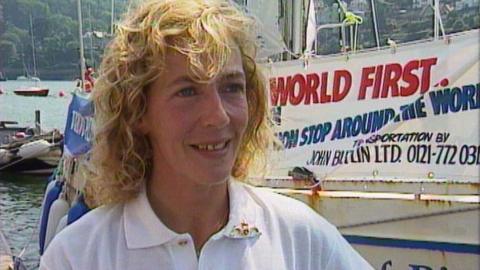 Woman with curly blond hair smiles. She is wearing a white polo t-shirt. She stands beside boats and the sea. 