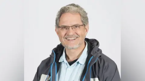 Councillor Brian Wernham. He has short grey curly hair and trimmed beard. He wears glasses, a light blue shirt and and outdoor grey and blue jacket. The head-and-shoulders portrait was taken against a white background. 