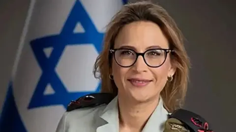 IDF handout A handout image from the Israel Defense Forces shows a head shot of former Israeli Military Advocate General Yifat Tomer-Yerushalmi. She wears glasses and is smiling at the camera, with an Israeli flag visible in the background. 