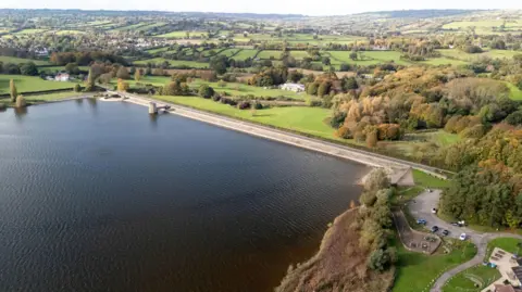 Getty/Colin Peachy Aerial, drone footage of the lake from the air. There is a road where the lake ends with the dam. There are lots of green fields and farmland in the background.