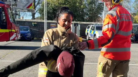 A woman dragging a dummy, with a firefighter wearing an orange jacket next to her.
