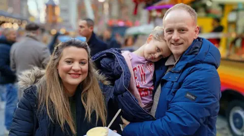 BBC Rebecca Stacey holds a pot of apple pie and custard as her husband holds her four-year-old daughter