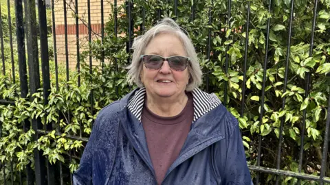 Liz McKenzie, a mature lady with a grey bob hairstyle and patterned glasses, stands in front of a fence with greenery behind it. She wears a purple T-shirt and a navy rain jacket which is wet from the rain.