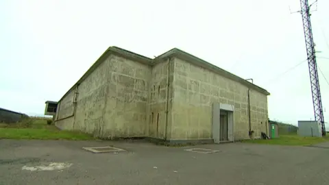 Exterior of former nuclear bunker. The stone building is grey. A door out of the building leads to a grass area and a tarmac area. A mast is next to the building. 