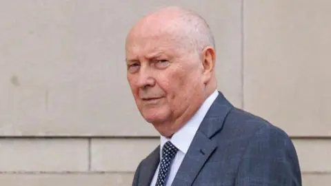 Frank Cushnahan, pictured outside Belfast Crown Court, has thin grey hair and is wearing a grey suit jacket, white shirt and a navy polka dot tie.