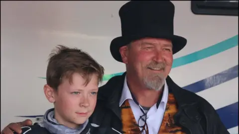 SB Media Solutions A young Storm Stacey, approximately 12 or 13 years of age, dressed in a black, grey and white race suit. He's standing next to his dad and having their photograph taken. Storm's dad is wearing a black top hat. He has a grey goatee type beard and smiling, holding Storm with his arm around his shoulder. He's wearing a patterned golden/orange waistcoat with a white shirt underneath and a black coat over the top.