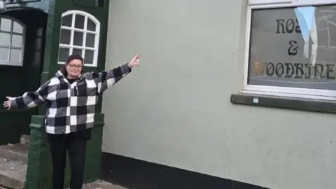 A woman standing in front of the entrance to a pub. She is wearing a black and white coat and black trousers. She has her arms out to the sides as she is celebrating becoming the pub's landlady.