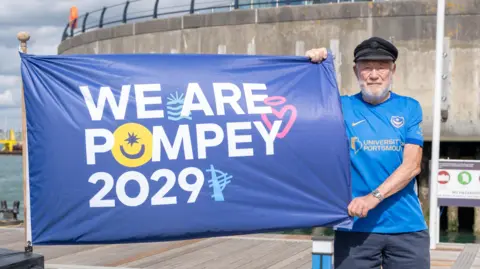 A man wearing a black hat and a blue t-shirt which reads 'University of Portsmouth stands to the right of the picture holding the corners of a navy blue flag. The flag reads 'WE ARE POMPEY 2029' in white letters.