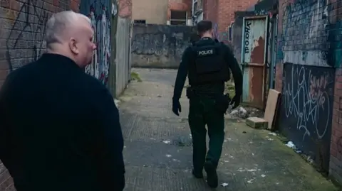 BBC/ Third Street Studios Two people walking up an alley with their backs to the camera. One closer to the camera is wearing a navy jacket and has white hair. The one further up the alley is a police officer wearing uniform that is black and dark green, he has brown hair.