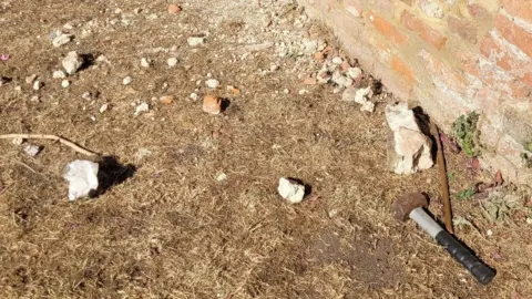 A close up of the base of the damaged brick wall. There is a hammer on the ground that it is believed was used to hit the wall. 
