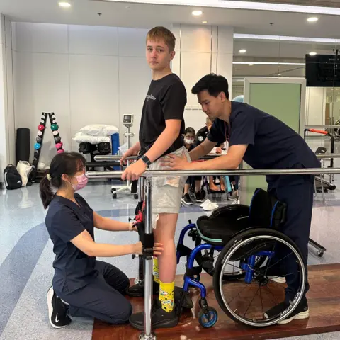 Kamila Nowicka A teenager is standing holding onto a bar with his wheelchair behind him. Two medical professionals are helping him.