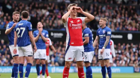 Reuters Arsenal's Leandro Trossard in red and white kit makes a binoculars gesture in celebration as a number of Ipswich players, wearing blue and white, speak to each other behind him.