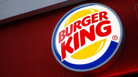 The image shows the Burger King logo on a big sign, which consists of "Burger King" written in big red letters with two burger buns on top and under the words. There is a blue ring surrounding it all, with white in the background.
The sign is on a red building.