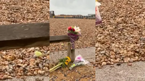 Flowers laid against a wooden post on a shingle beach.