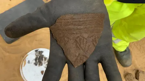 MOLA-Wessex Archaeology The gloved hand of an archaeologist holding a shard of decorated prehistoric pottery.