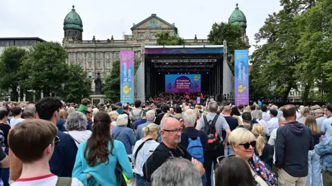 Pacemaker The image shows a brightly lit stage in the distances and a large crowd of people gathered in front. They have their backs to the camera. 