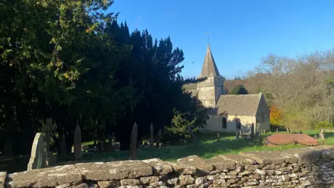 BBC The exterior of St Kenelm's in Sapperton. A small church with a pointed cone-shaped spire is visible in the background, with the graveyard in the foreground. 