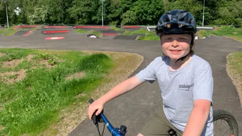 A young boy on a bike with a helmet on and a grey T-shirt stands in front of a bike track with numerous bumps and bends