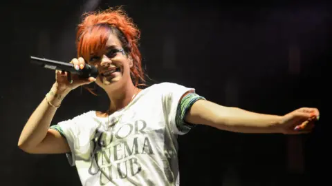 Andy Sheppard/Redferns via Getty Images Lily Allen performing onstage. She is smiling and singing into a microphone while wearing a Two Door Cinema Club T-shirt.
