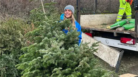 Shooting Star Children's Hospices A woman with long, blonde hair wearing a grey beanie and blue coat standing behind a Christmas tree.