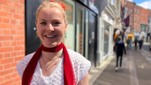 A woman has blonde hair, scraped back from her face in a red bobble. She is smiling into the camera. She wears a white top and a skinny red scarf.