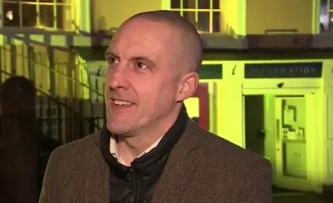 Gavin Capstick speaking to a reporter out of view outside of the Carlisle tourist information point. He has a shaved head and brown eyes. He is wearing a brown coat and black jumper.