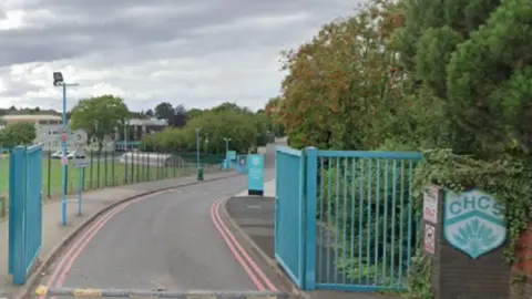 Blue gates stand at a long concrete driveway entrance to the school with a building far off in the distance and grass playing fields off to the left. A sign saying CHCS is on a wall to the right of the entrance with ivy and greenery growing above and around it.