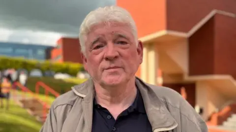 Joseph is looking at the camera. He has a plain expression and has white hair. He is wearing a beige zip up jacket and a navy polo top.