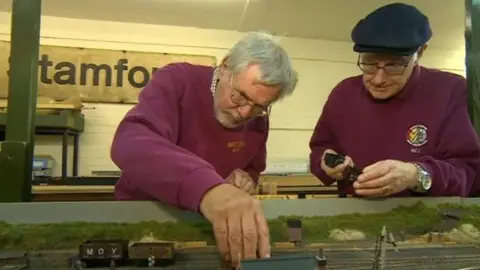 BBC Two men in burgundy jumpers are leaning over a model railway display intently as they check the track and hold black items to go on it. A workshop can be seen on the wall behind them.