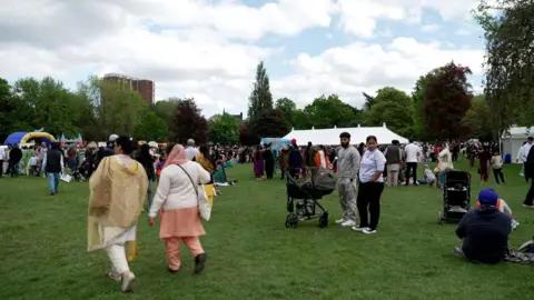Crowds of people walking through a park. A white tent is in the middle of the grass and a blue bouncy castle is in the far right of the image.