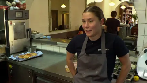 Sasha, a young woman in a pub kitchen. She has an apron on over her black t shirt and her hair is tied back