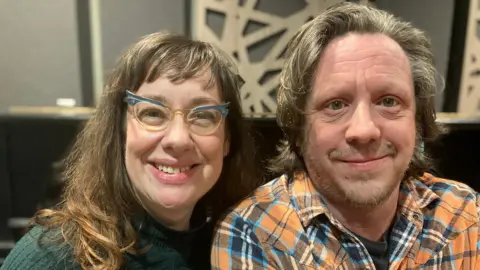 The picture shows a woman on the left with long brown hair and green rimmed glasses and a green jumper siting next to a man with grey brown hair and an orange checked shirt. Both are looking at the camera and smiling.