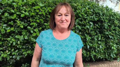 Tricia Timms wearing a blue and white patterned top and standing in front of a holly bush. She has shoulder-length brown hair with a slight fringe, and is smiling at the camera with her arms down beside her.