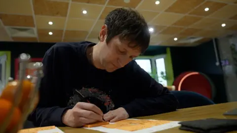 Tim Everest/BBC A man writing on orange sticky notes, some of which are arranged on a sheet of paper on the desk in front of him.