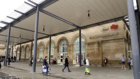 The exterior of Hull's Paragon Station. People are walking by the yellow stone building, which has several large glass doors at its entrance