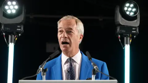 Nigel Farage in a bright blue suit, white shirt and black tie, with red and white coloured dots stands behind a lectern. Two lights are either side of him.