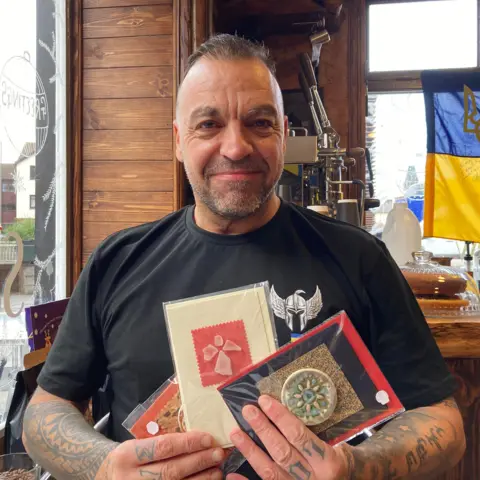 A man with short dark hair and tattoos on his arms wearing a black t-shirt holds a set of Christmas cards while smiling at the camera.