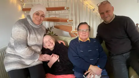 Pictured is Bushra, Margaret, George and Khaled all sat on a white leather sofa in the living room. Bushra is wearing a nude headscarf with pearl embellishments attached on it, a high neck wool-like jumper that is grey, brown and beige. Margaret is wearing a black jumper and pink trousers. She has her head laid on Bushra and is smiling. George wears a yellow collar shirt underneath a blue jumper and has slicked-back brown hair. Khaled has his arm wrapped behind George's shoulder and is smiling. He wears a dark brown jumper with a beige collar shirt underneath and dark blue jeans with his black framed glasses sitting on the top of his head.
