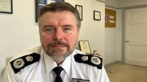 Chief constable Tim Smith is in an office at Kent Police with framed certificates on the wall behind him and the force logo. He is wearing his uniform and has grey hair and a beard.