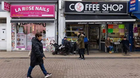 A woman in a black coat walks along a high street in Walthamstow, passed a Coffee shop and a nail salon with bright pink signage.