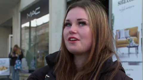 A woman, with shoulder length brown hair, looking at the camera. She is wearing a black coat. Behind her is House of Fraser. 
